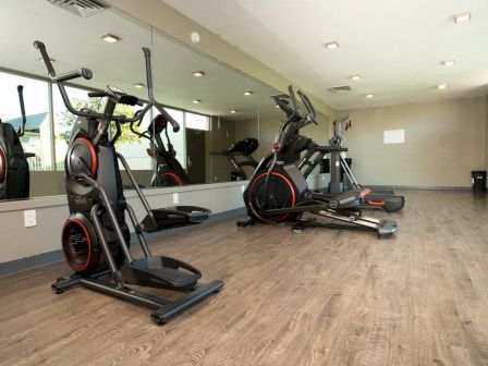 A gym room with exercise bikes and equipment on a wooden floor, mirrored wall reflecting the machines, and overhead lighting.