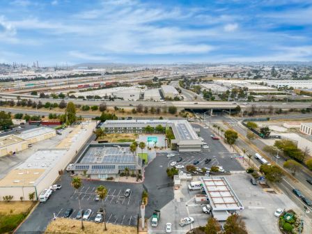 The image shows an aerial view of an industrial and commercial area with roads, buildings, a pool, and parked cars under a partly cloudy sky.