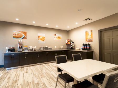 A small breakfast buffet area with coffee machines, cereal dispensers, and seating. Wall art features breakfast foods.