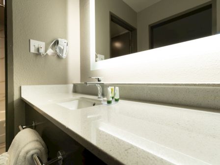 A modern bathroom vanity with a sink, mirror, two small toiletries, mounted hairdryer, and folded towel; bathtub seen through the doorway.