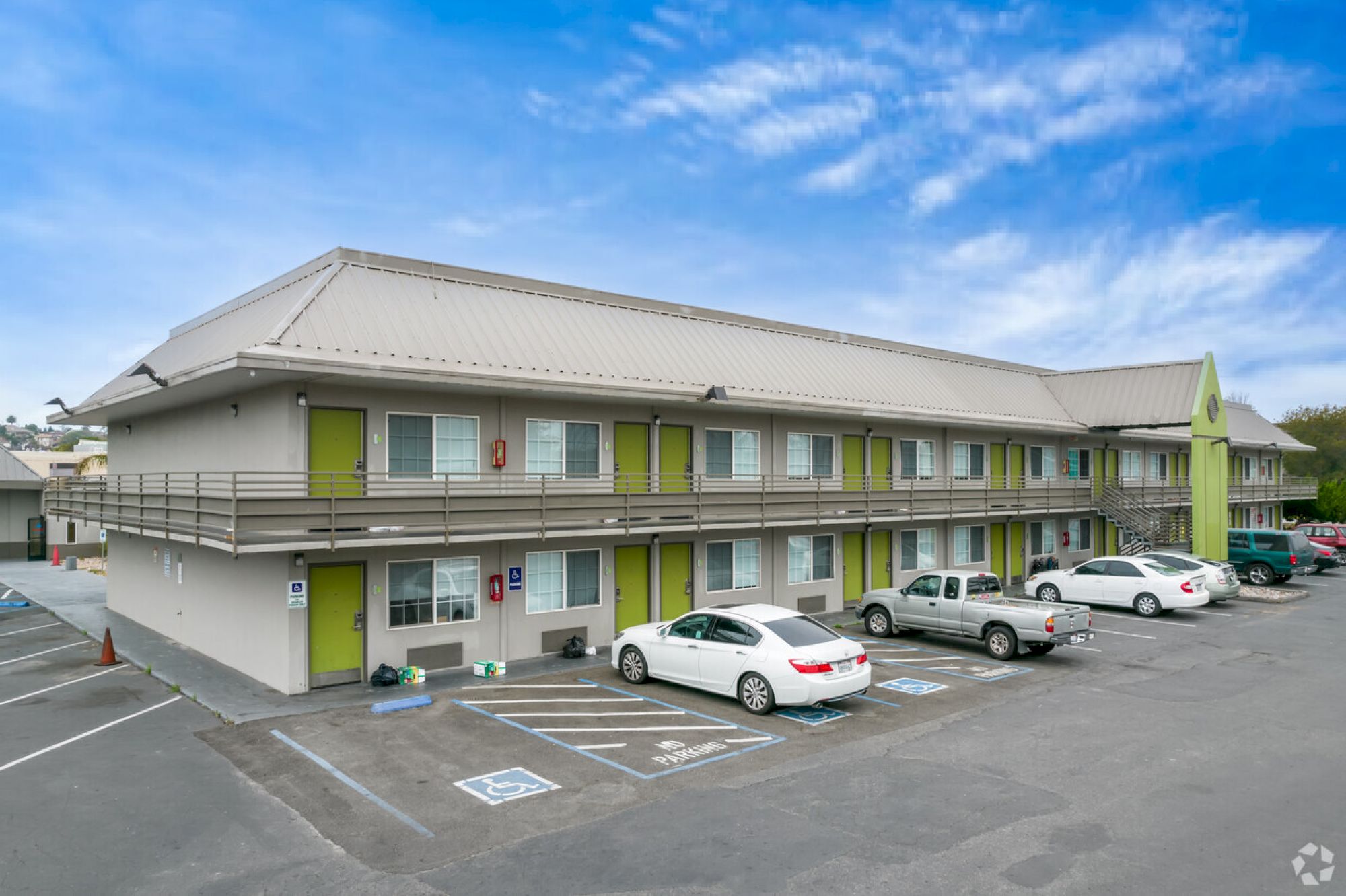 The image shows a two-story motel building with several parked cars in front, featuring green accents and a blue sky background.