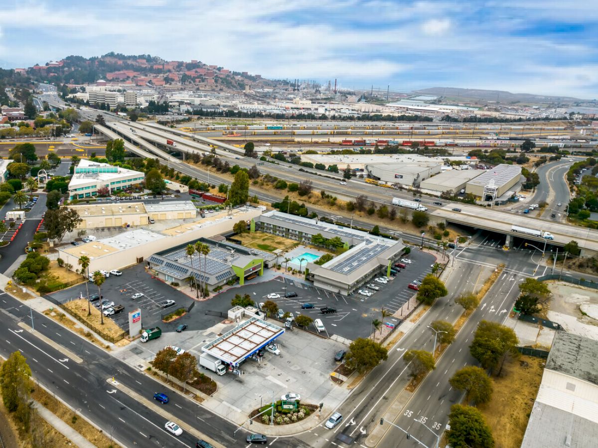 The image shows an aerial view of a commercial area with roads, buildings, and a gas station, set against a backdrop of hills and rail tracks.