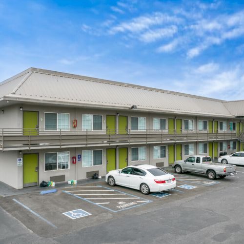 A two-story motel building with green accents and a parking lot with several cars in front, set against a blue sky.