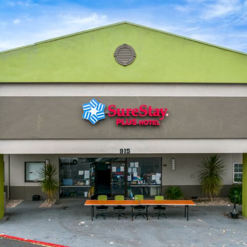 The image shows the entrance of a SureStay Plus Hotel building with green accents, tables, chairs, and a clear blue sky above.