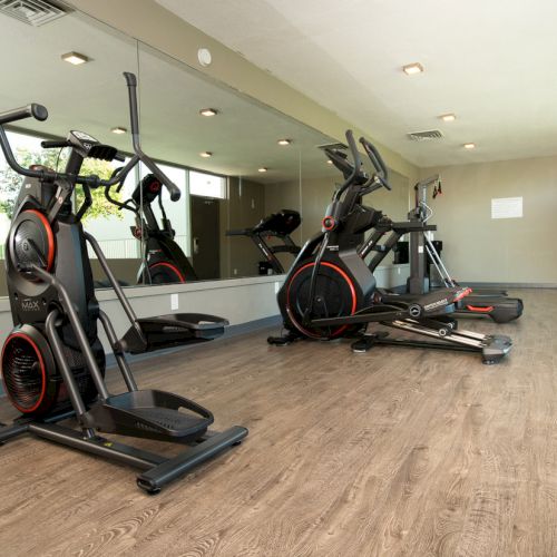 A gym room with two elliptical machines on a wooden floor, mirrors reflecting the equipment on the wall and recessed lighting above.