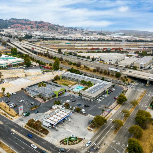 Aerial view of a commercial area with roads, buildings, and a gas station. Hills and industrial infrastructure in the background.