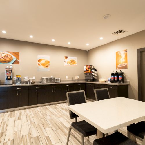 A small breakfast area with tables, chairs, a counter with coffee machines, waffle makers, and cereal dispensers. Warm lighting and decor.