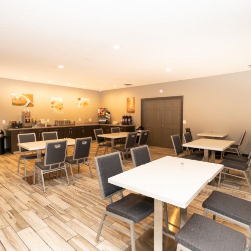 The image shows a dining room with tables, chairs, and a counter area with drink dispensers and food items at the back.