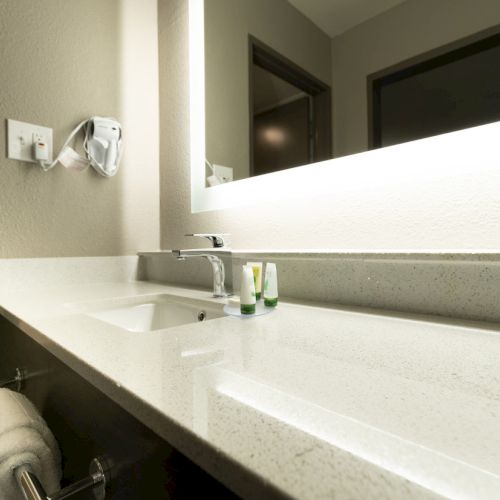 A modern bathroom vanity with a sink, backlit mirror, a mounted hairdryer, towels, and toiletries on the counter.