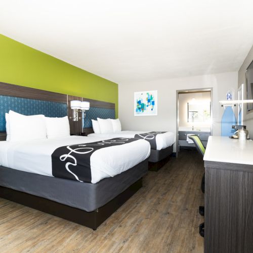 A modern hotel room features two double beds, a flat-screen TV, a desk, and bright green accent wall, creating a welcoming atmosphere.