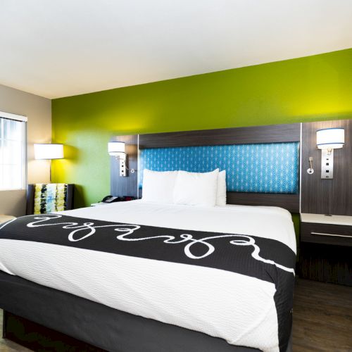A modern hotel room with a large bed, two bedside tables, wall lamps, a green accent wall, window, and framed artwork.