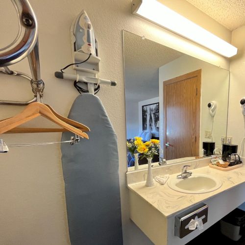 A bathroom with a mirror, sink, ironing board, hairdryers, and a vase of flowers on the countertop beside toiletries.