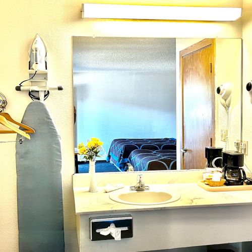 A hotel vanity with a sink, mirror, iron, flowers, hangers, coffee maker, and hairdryer; a glimpse of a bedroom in the mirror.