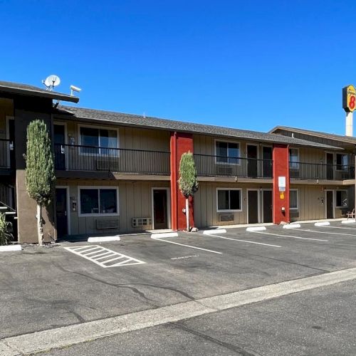 The image shows a two-story motel with a parking lot, featuring colorful pillars and a visible motel sign against a clear sky.
