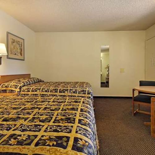 The image shows a hotel room with two beds, a small table with chairs, a lamp, a wall mirror, and a window.