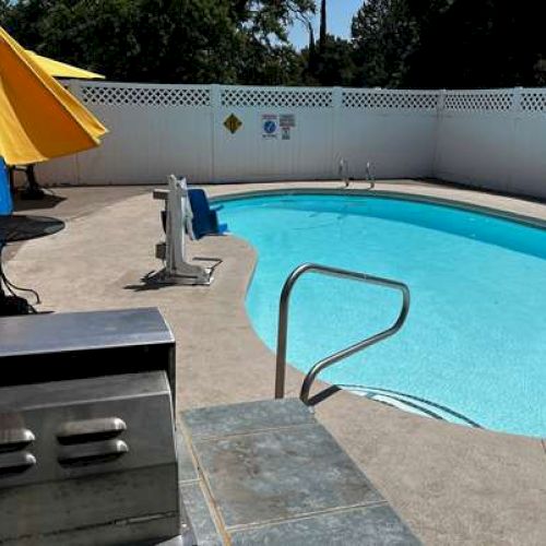 A small outdoor pool with a metal railing, a barbecue grill, and a yellow umbrella providing shade over a sitting area nearby on a sunny day.