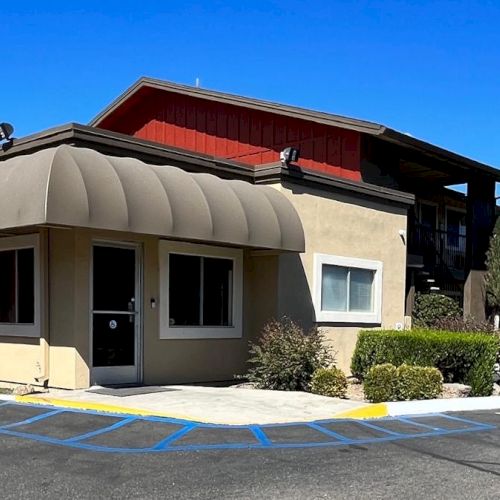 The image shows a motel with a beige and red exterior, surrounded by a parking lot and some greenery, under a clear blue sky.