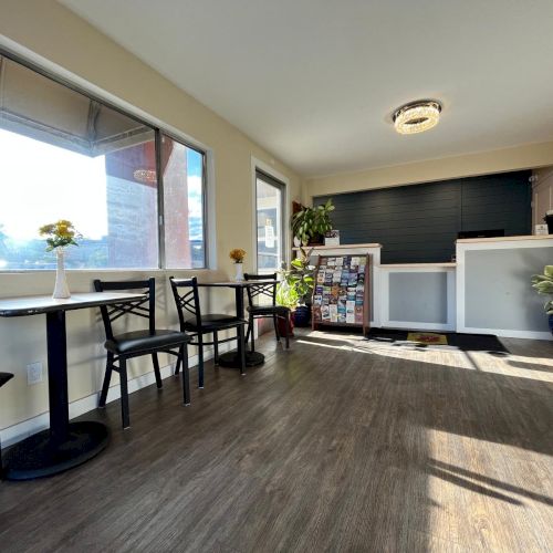 A reception area with tables, chairs, potted plants, and a counter. Sunlight streams through large windows creating a bright atmosphere.