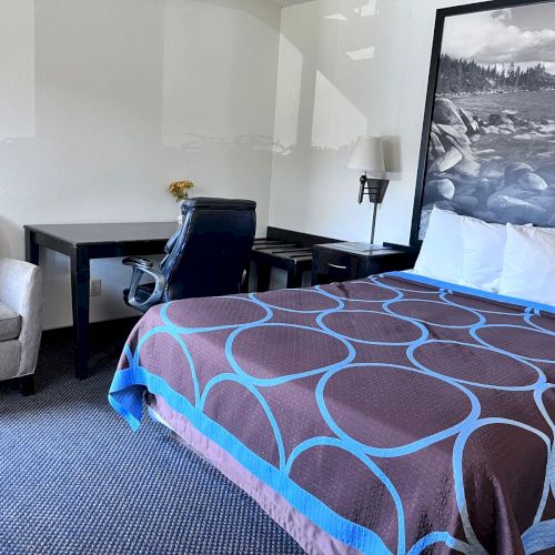 A hotel room with a bed, a blue and brown-patterned blanket, a desk with a chair, a sofa, and a large black-and-white wall picture.