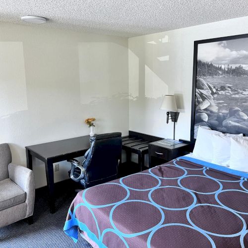 A hotel room featuring a bed, chair, desk with chair, wall art, and a lamp, creating a neat and cozy ambiance.