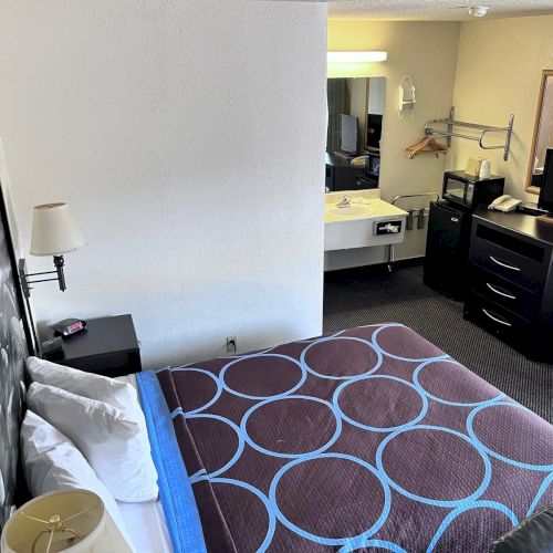 A hotel room features a bed, nightstand, lamp, television, dresser, and a sink area with a mirror and hangers on the wall.