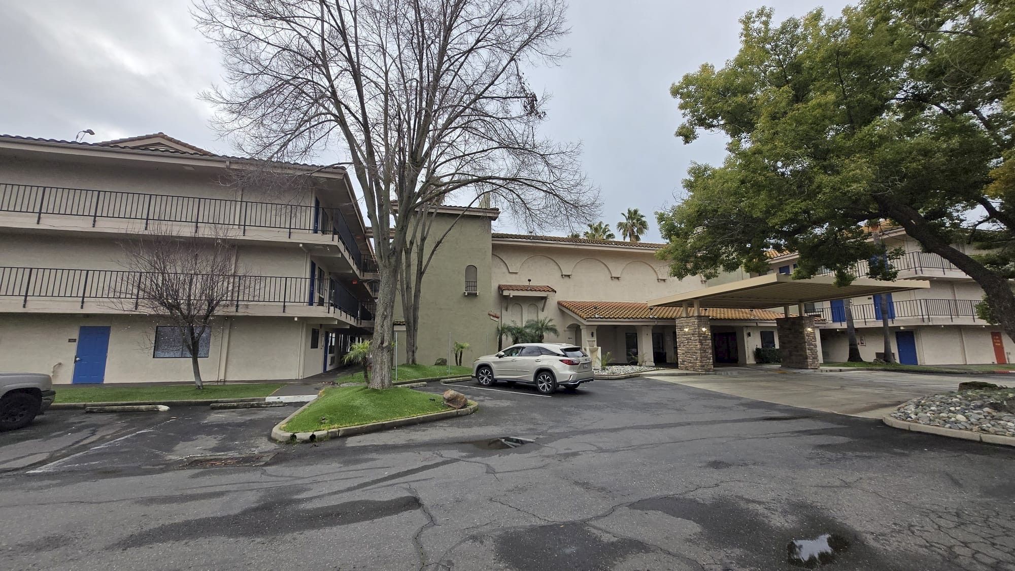 The image shows a multi-story building with balconies, a tree, and a parked white car, set against a cloudy sky.