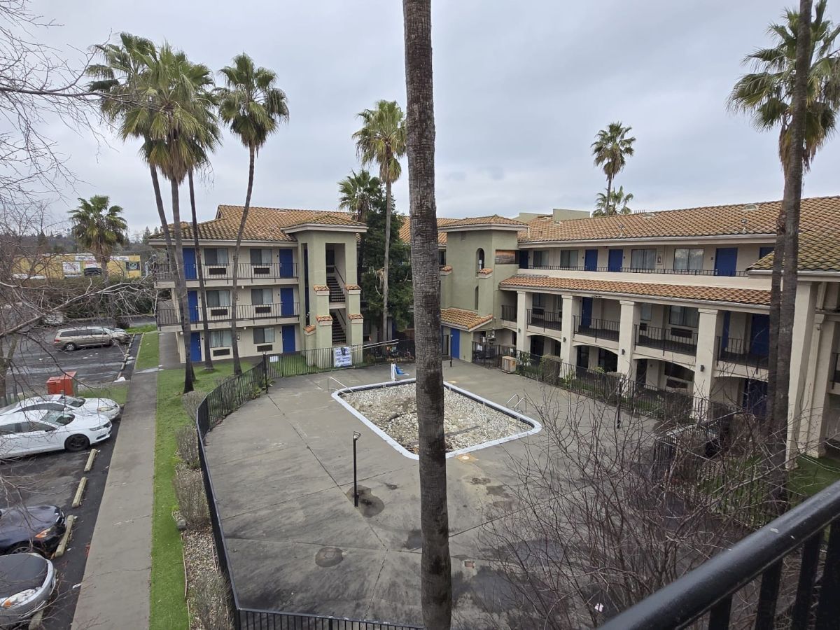 The image shows a multi-story hotel or apartment building with an empty pool, surrounded by palm trees and parked cars on a cloudy day.