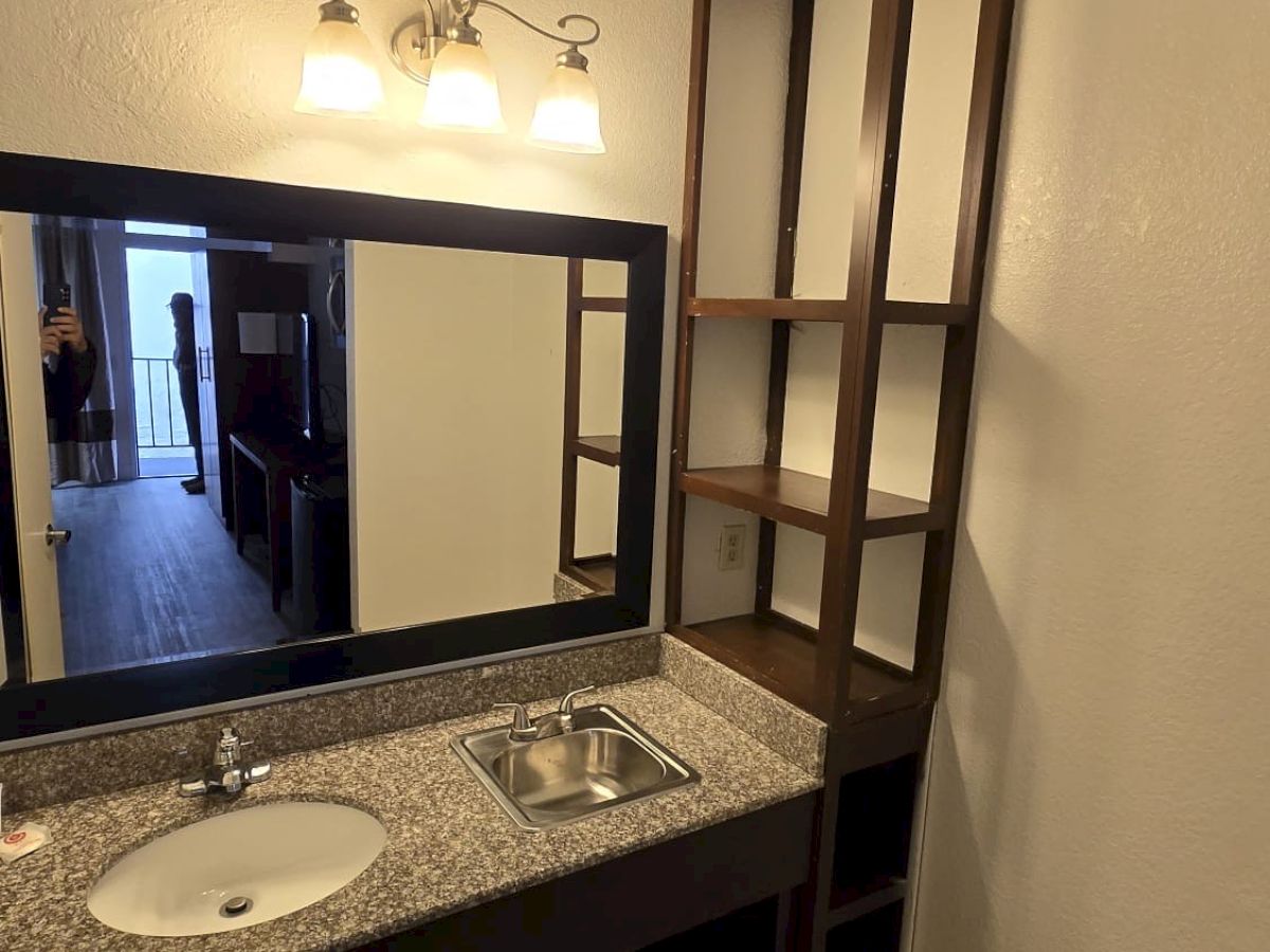 A bathroom with a sink, granite countertop, mirror, wall lights, and tall shelving unit. Floor is tiled, and a person is visible in the mirror.