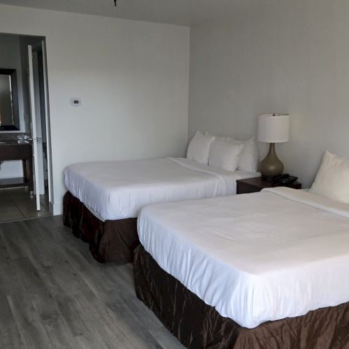 A hotel room with two double beds, a TV, a lamp, and open view to a bathroom area with a sink and mirror.