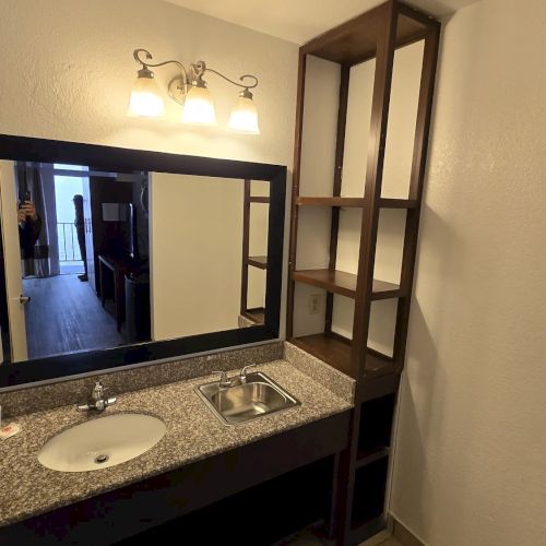 A bathroom with a large mirror, sink, countertop, and shelves on the side, illuminated by wall-mounted lights.