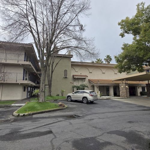 The image shows a three-story building with balconies, trees, and a white car parked in front, set against a cloudy sky.
