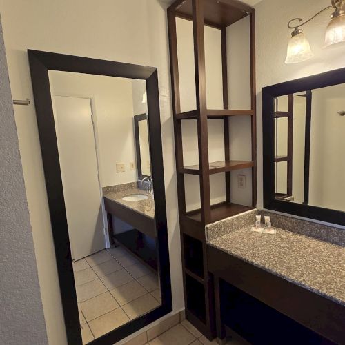 A bathroom with a granite countertop, sink, mirror, shelving, and a door leading to another room is pictured.