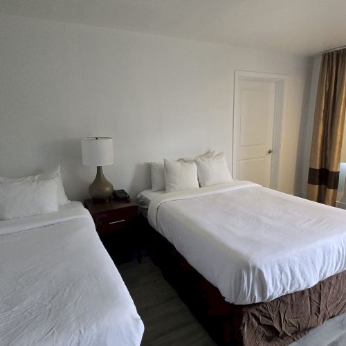 The image shows a hotel room with two beds, a nightstand with a lamp, curtains over a window, and an air conditioning unit below.