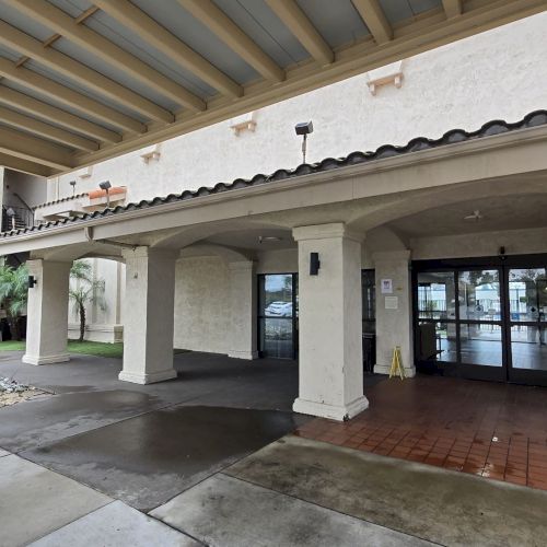 An exterior view of a building entrance with pillars, a covered walkway, tiled floor, and wet pavement, suggesting recent rain.