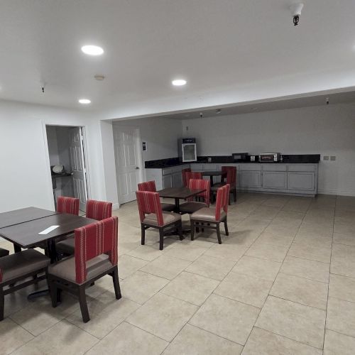 The image shows a room with tables, chairs, a lamp, and a kitchenette, likely a communal space or dining area with tiled flooring.
