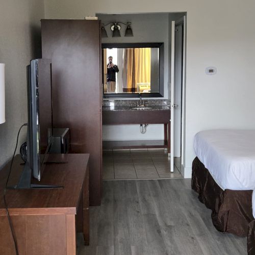 The image shows a hotel room with a bed, a TV on a wooden desk, a mirror, a sink, and laminate flooring.