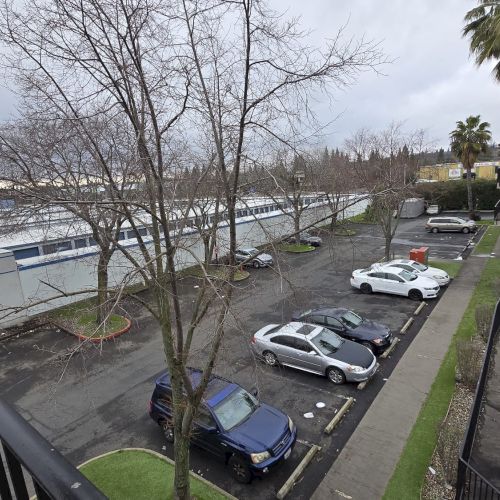 A parking lot with several cars, surrounding trees, a large building on the left, and a hotel or apartment complex on the right.