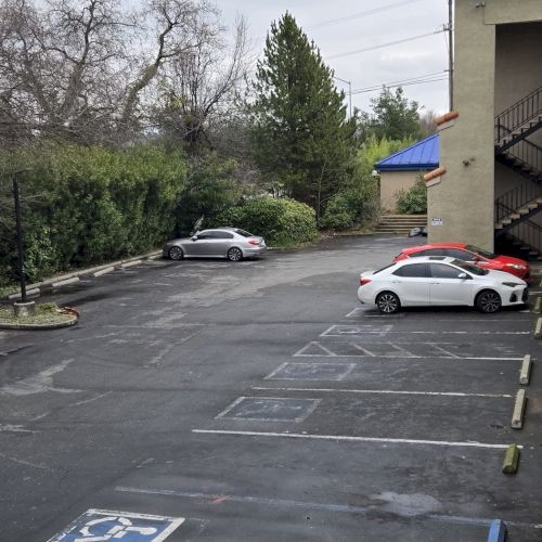 A parking lot with a few cars, including a white and a red one, next to a building with stairs and trees in the background.