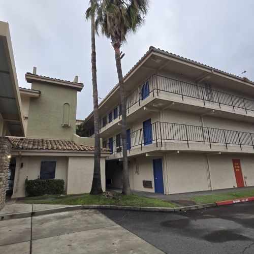 The image shows a three-story motel building with external corridors, surrounded by palm trees, with cloudy skies above.