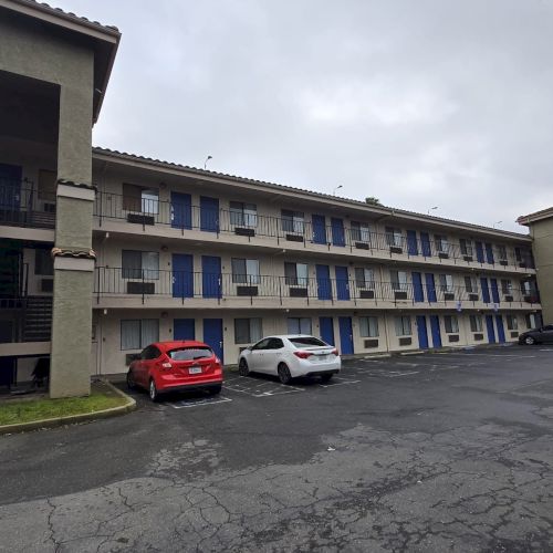 A three-story building with exterior corridors, parked cars, and palm trees on a cloudy day.