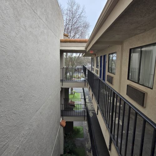 A narrow alleyway between two buildings with balconies and glass windows, leading to an outdoor area with greenery and visible sky above.
