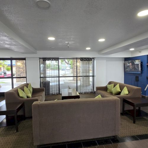 A modern lobby with sofas, green cushions, a TV, and coffee area. Large windows and ceiling lights provide a bright atmosphere.