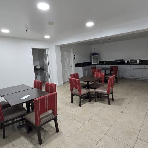 A spacious room with several tables and red chairs, a kitchen area with appliances, and tiled floor. The lighting is bright and even.