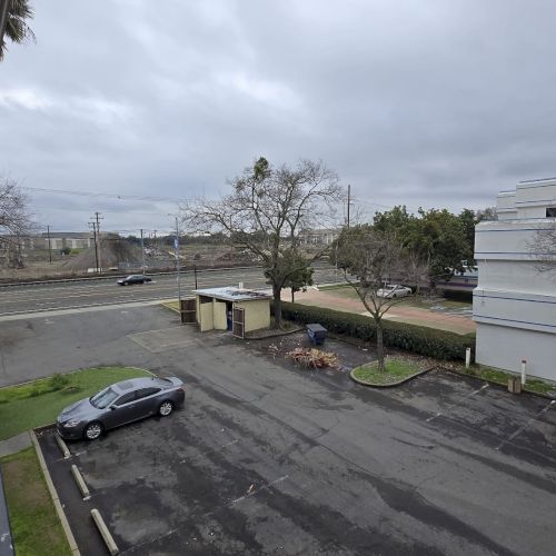 A parking area with a silver car, trees, and a white building. A road and distant landscape are visible under an overcast sky.