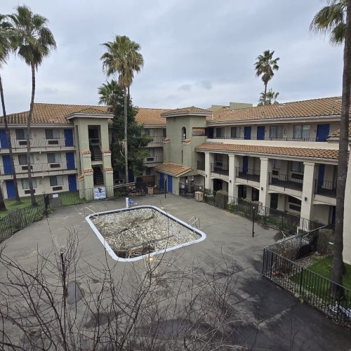 The image shows a three-story building with a courtyard and palm trees, parking on the side, and an empty rectangular pool in the center.