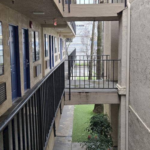 The image shows a view of a multi-level building's hallway with railings and doors, overlooking a small garden area below.