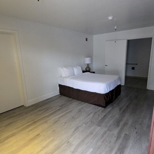 A minimalist room with a bed, side table, and lamp on a wooden floor, featuring white walls and a closed door on the left.