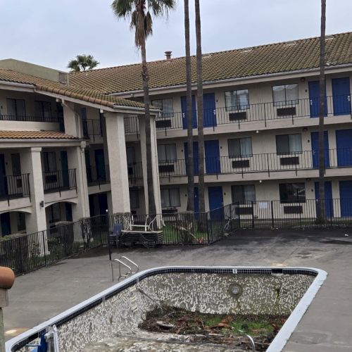 The image shows an empty, neglected swimming pool surrounded by a multi-story building with balconies and several palm trees in the background.