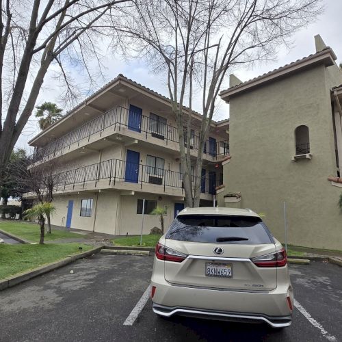 A parking lot with a white car, trees, and a beige multi-story building with blue doors. Overcast sky adds to the setting.
