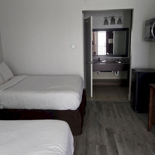The image shows a hotel room with two beds, a TV, a desk, and a lamp. A bathroom sink and mirror are visible through an open door.